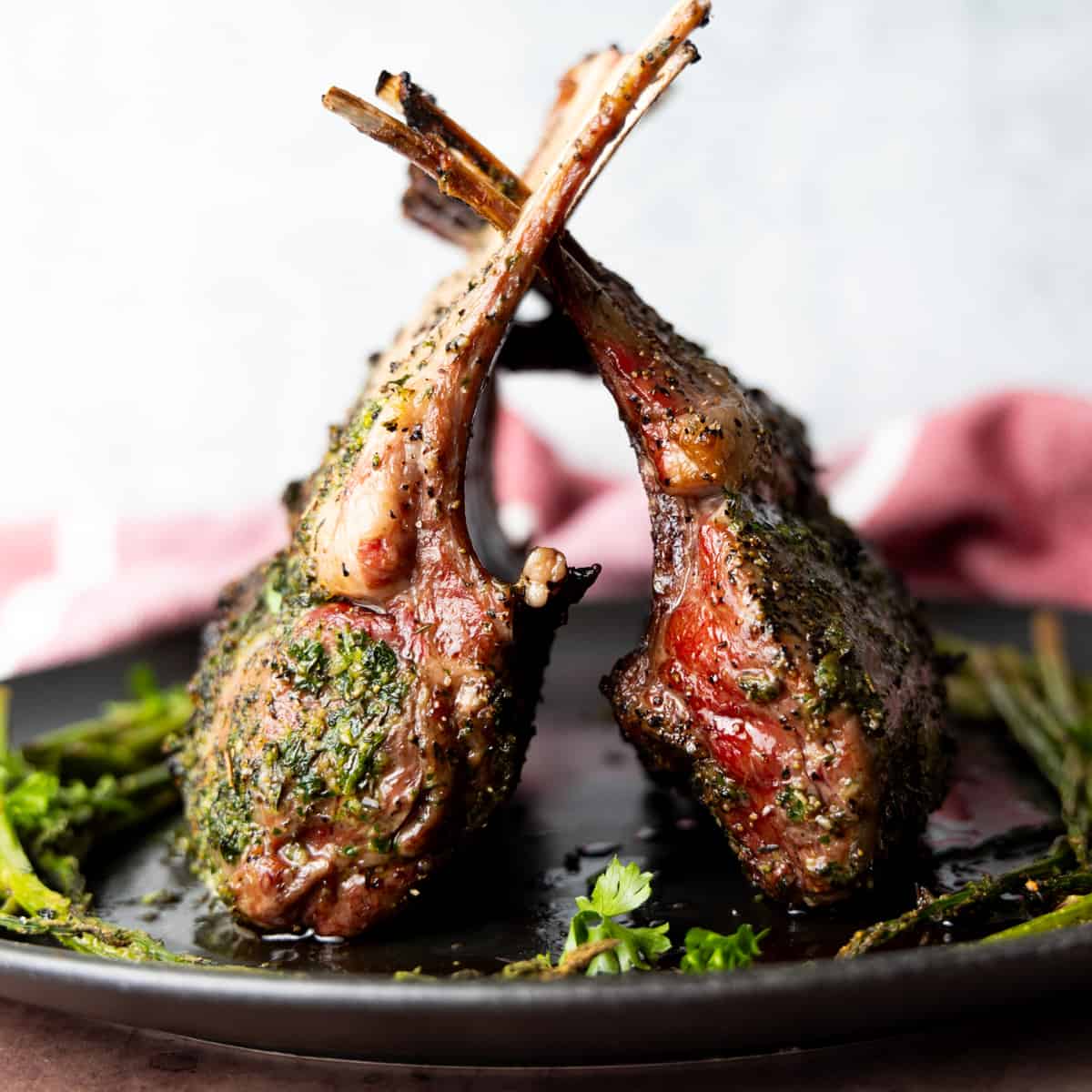 Two racks of lamb on black plate standing tall leaning on one another with asparagus surrounding meat.