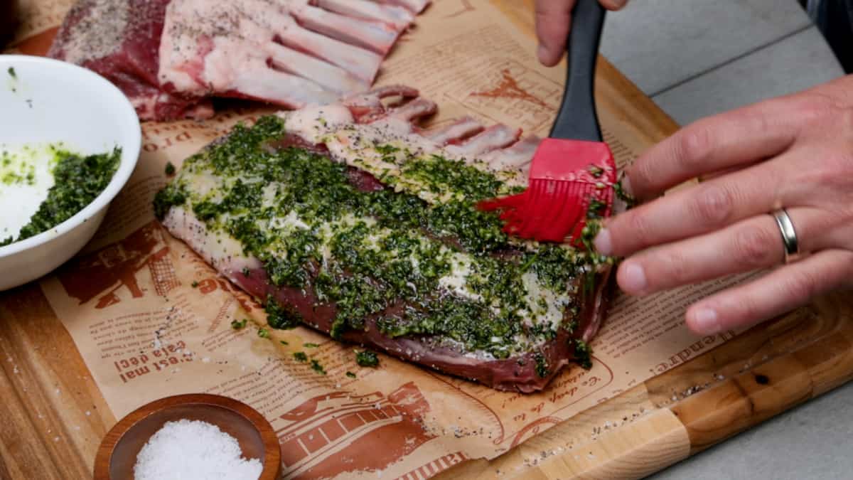 Adding marinade to lamb chops atop a wood cutting board by hand.