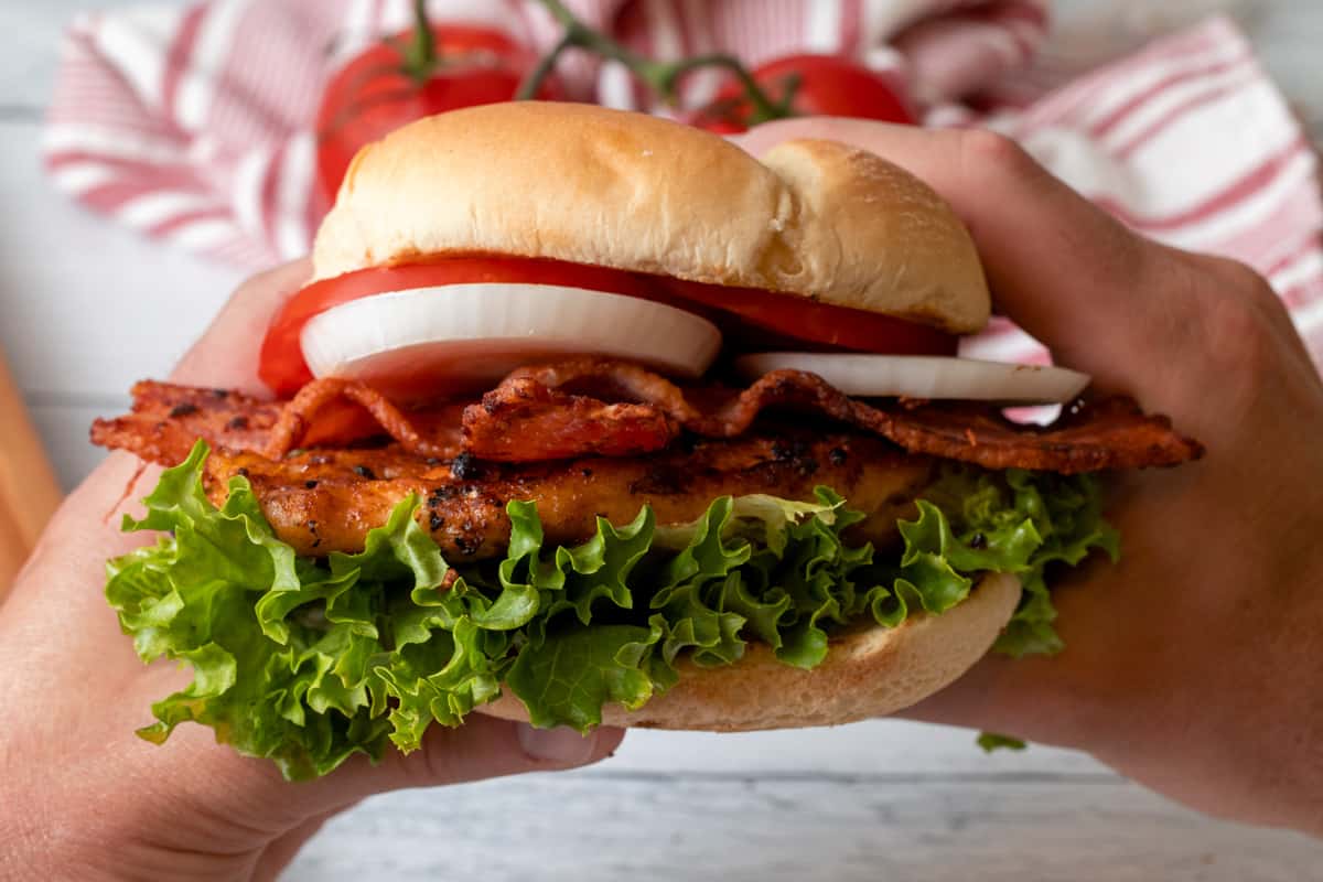 Chicken sandwich with lettuce, onion, tomatoes, and bacon being held with two hands.