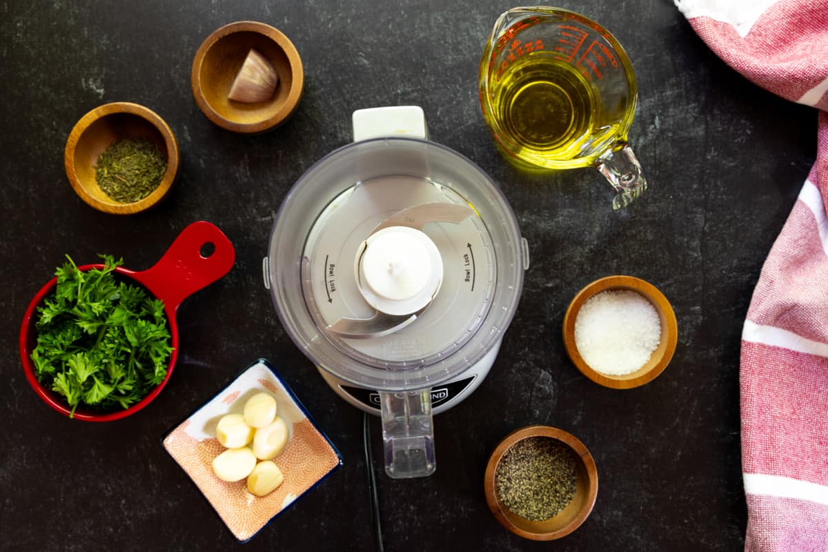 Food processor with small dishes of garlic, parsley, shallot, salt, pepper, & olive oil.