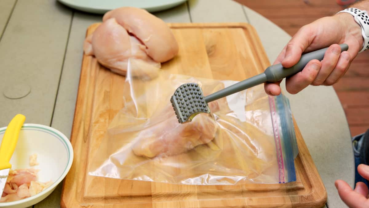 Chicken breasts in ziplock bag on cutting board with a meat mallet being held by hand over chicken.