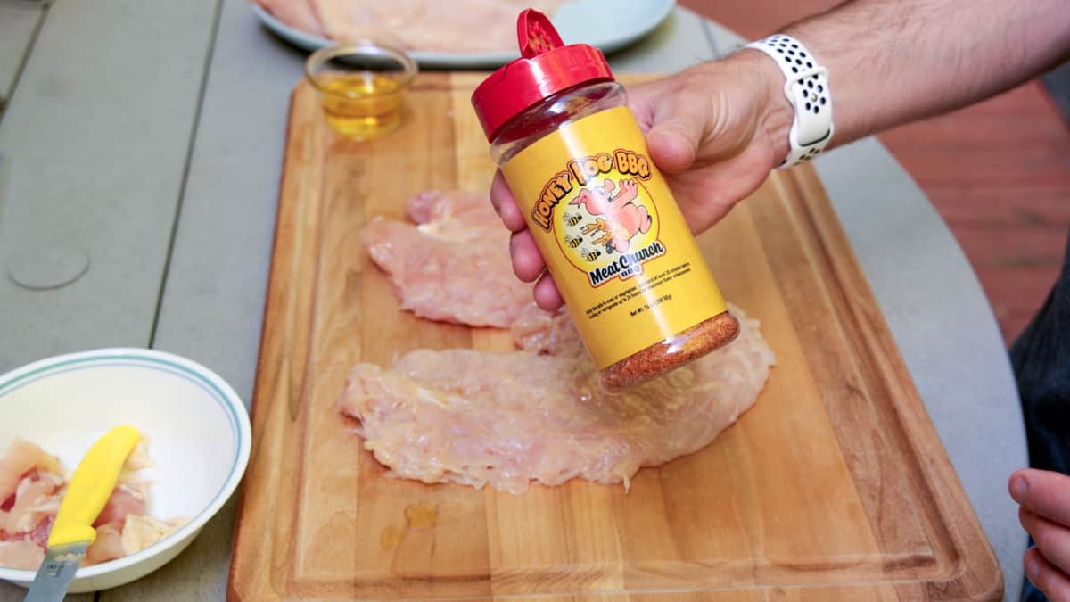 Chicken breasts on cutting board with a hand holding meat church brand seasoning in bottle. 
