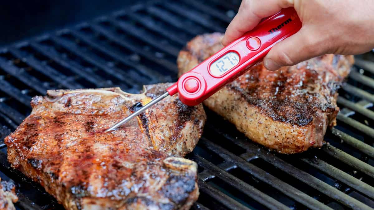 Steaks on grill being tested with meat thermometer.