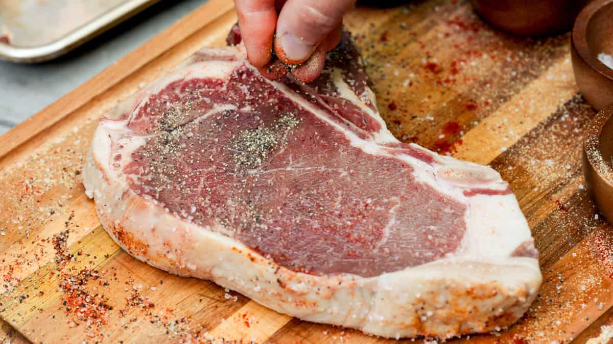 Seasoning a t bone steak on wood cutting board with salt and pepper.